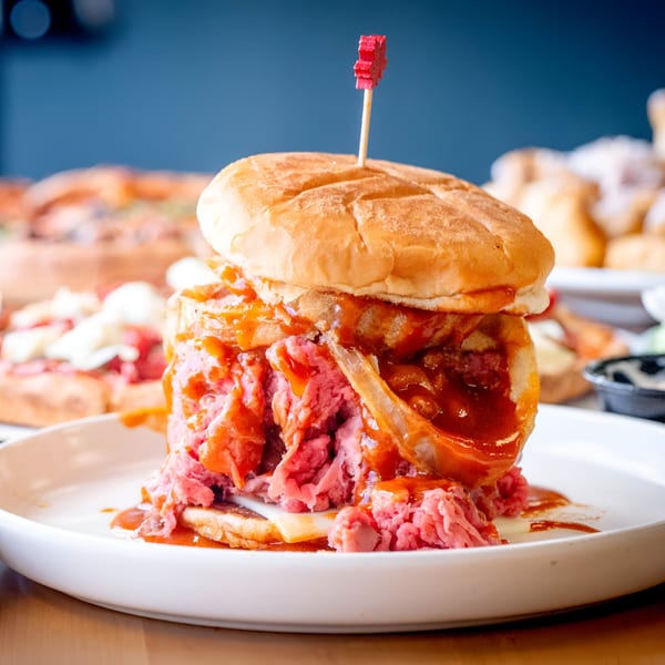 A towering Apollon Roast Beef sandwich with sauce, cheese, and a red pick on a white plate.