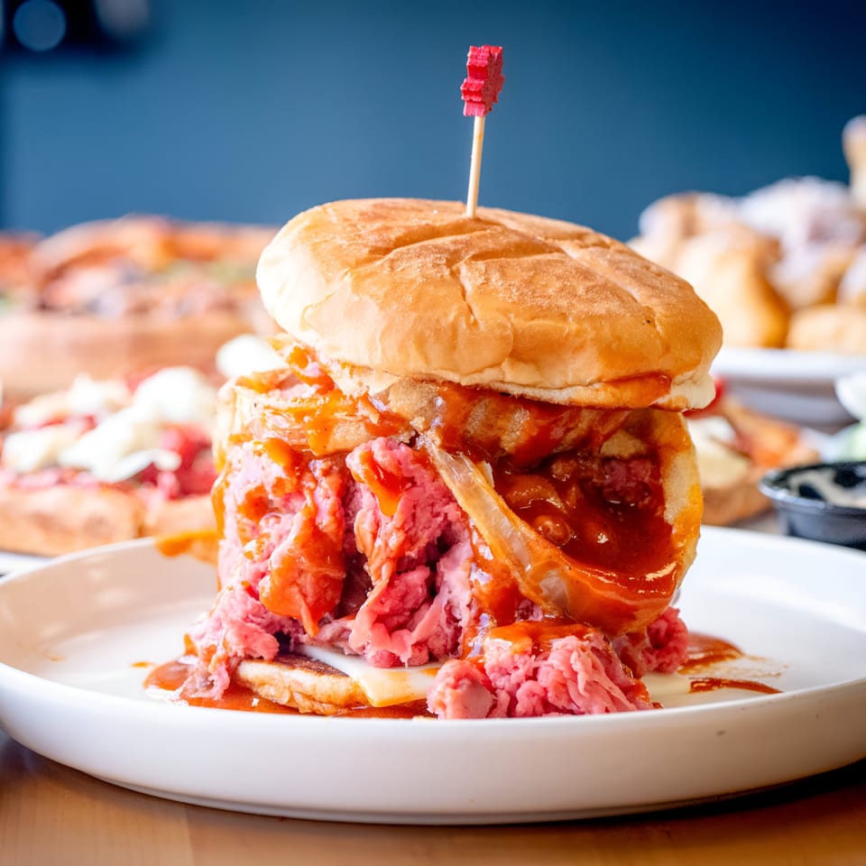 A towering Apollon Roast Beef sandwich with sauce, cheese, and a red pick on a white plate.